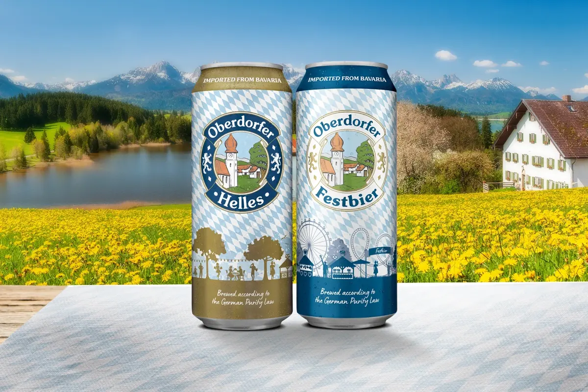 Two cans of Oberdorfer Helles and Oberdorfer Festbier on a table in front of a mountain landscape with a lake.