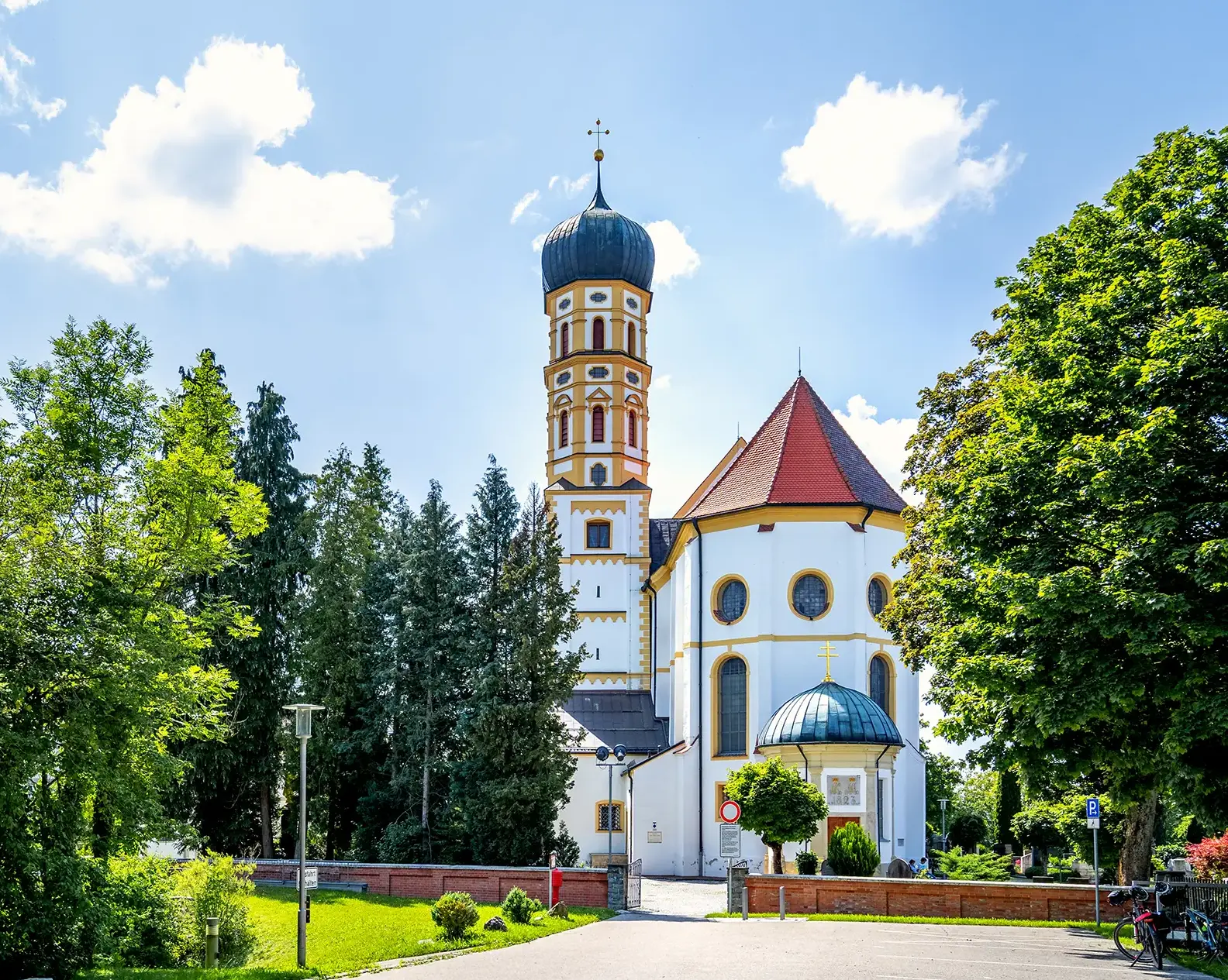 A picturesque church with a tall, colorful tower and a red-tiled roof stands among lush green trees under a blue sky.