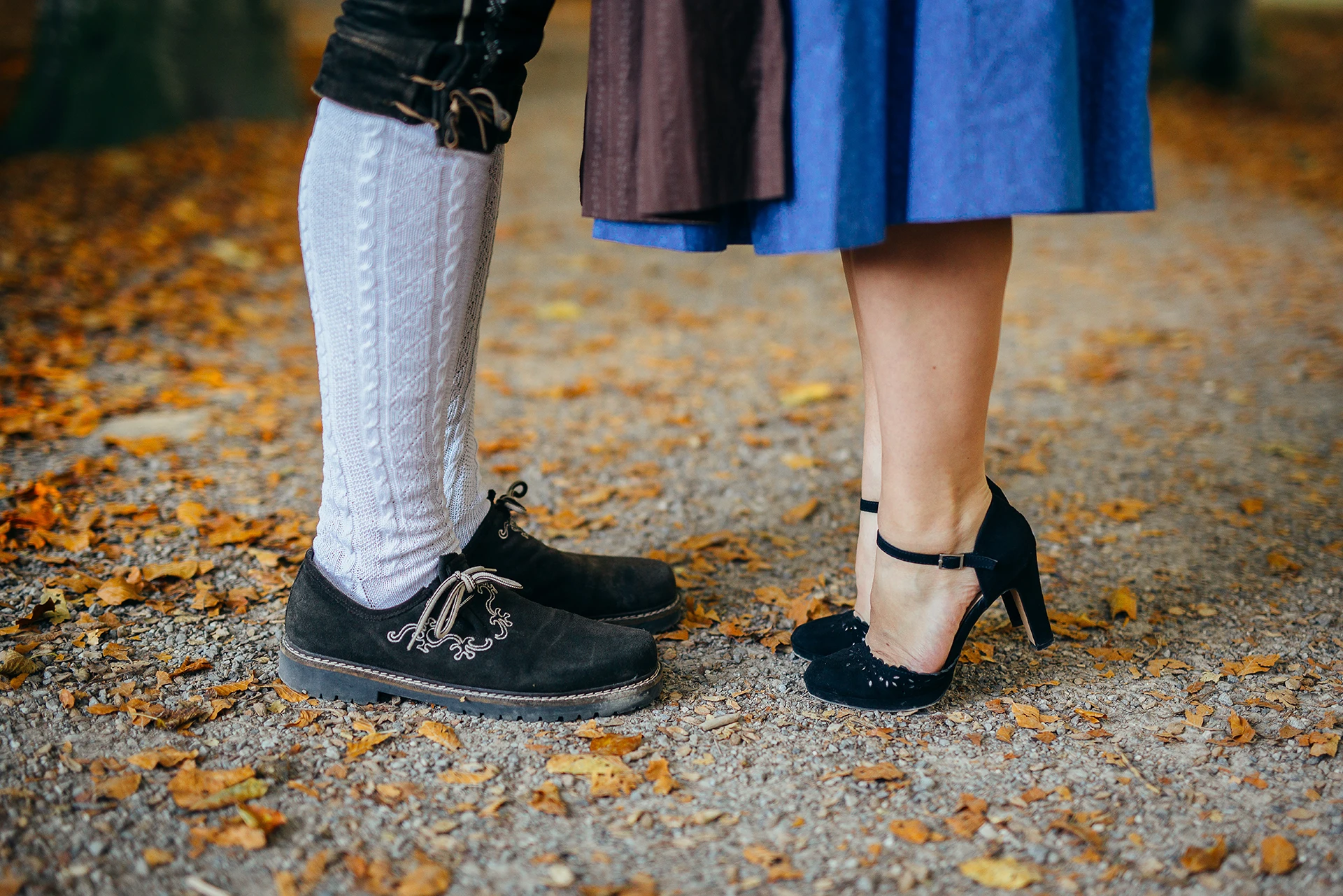 Two people stand close together, showing only from the knee down on a leaf-strewn path. One wears black suede boots and white knit leggings, the other black high heels.