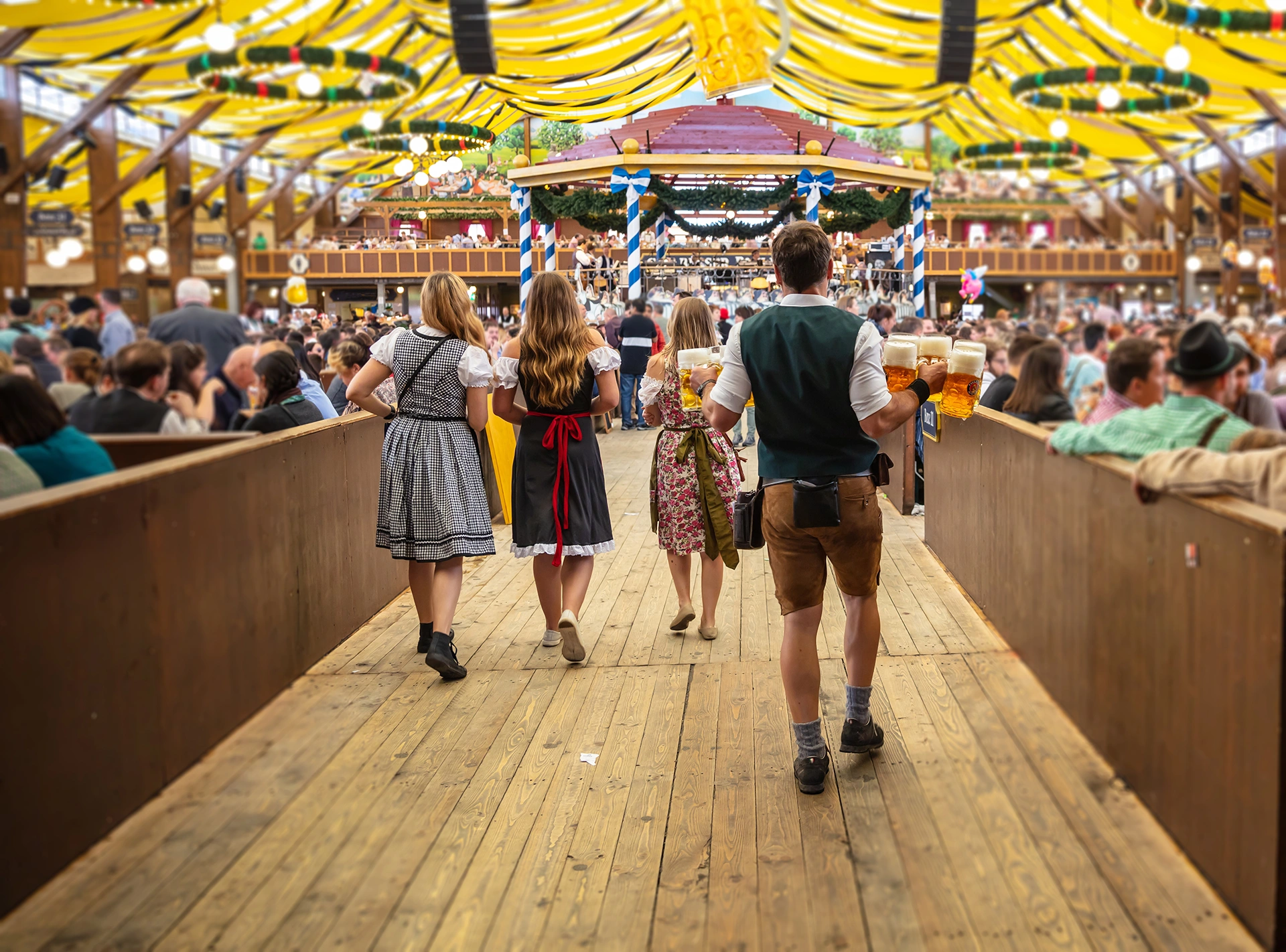 Several people in traditional dress are walking through a festival tent. A waiter is carrying several full beer mugs.