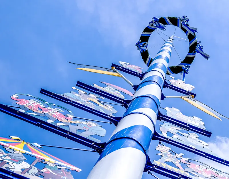 A maypole decorated with colorful, shield-shaped signs featuring medieval knights stands against a clear blue sky.
