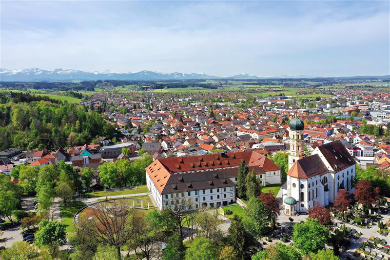 Aerial view of a quaint European town with a prominent church, surrounded by lush greenery and distant mountains.
